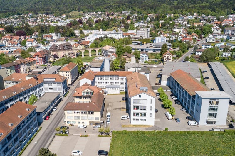 Building complex with multiple residential and commercial buildings, several cars parked in front, surrounded by greenery and a paved road.