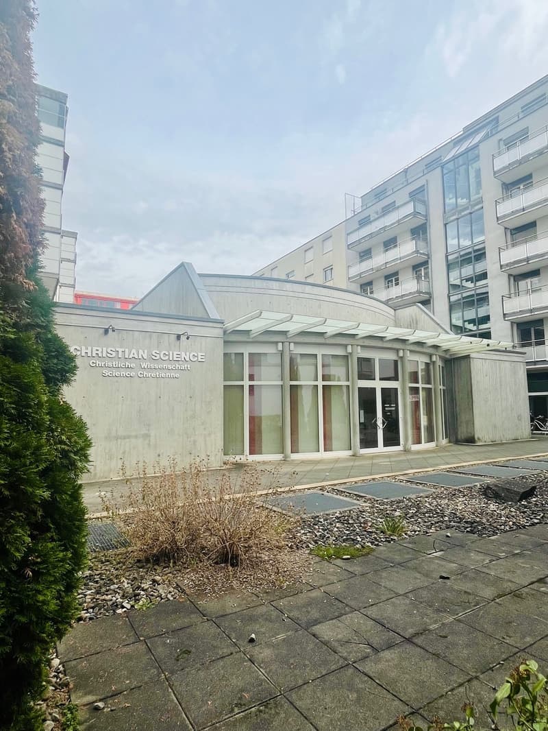 A modern building with gray concrete walls, glass doors and windows, and a small garden in front. The building is labeled as Christian Science. A tall building is on the right side. A bike is parked on the right.