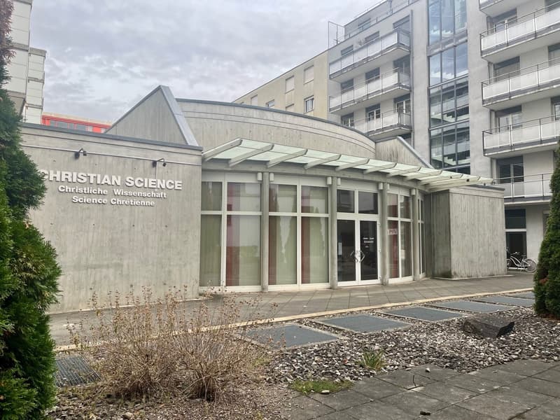 Building exterior of Christian Science with entrance, concrete wall, glass windows, glass doors, and bicycle parked outside.