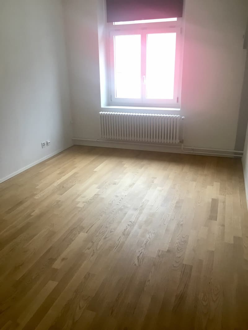 white walls, wooden floor, a window with white frames, white radiator, two white electrical outlets