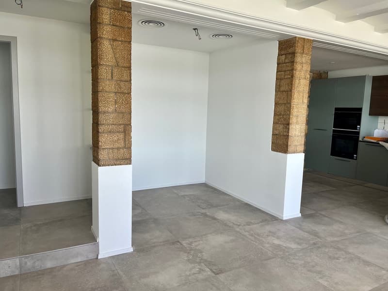 empty room with white walls, tile flooring, exposed brick columns, ceiling fixtures, doorway
