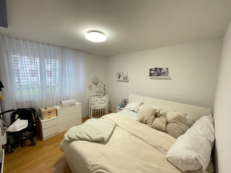 Bedroom with bed, crib, white walls, wooden floor, white curtains, white furniture, ceiling light, shelves with pictures and items