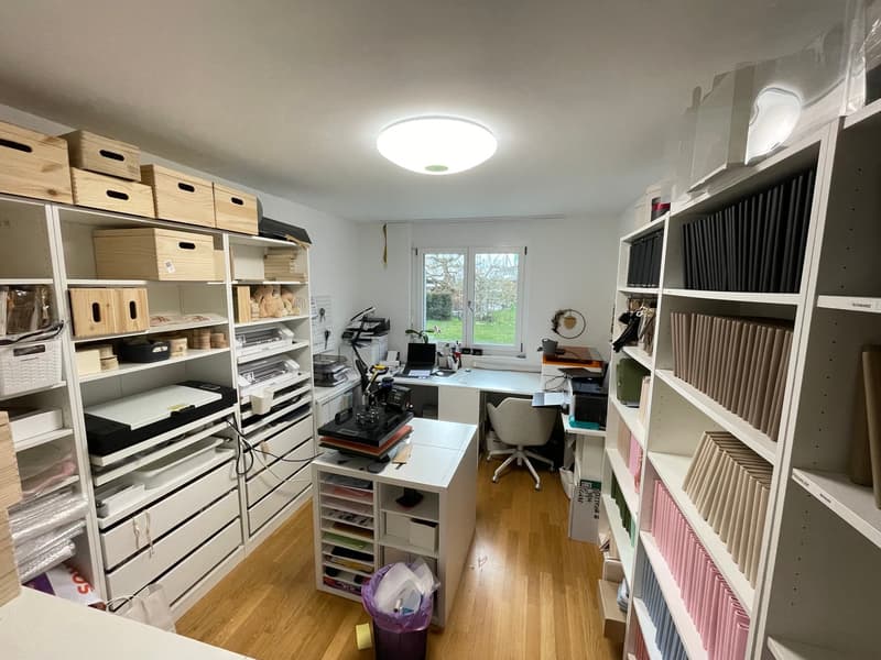 A room with wooden floor, white walls, shelves, desk, chair, printer, scanner, storage boxes, trash bin, and a window with a view of a grassy area.