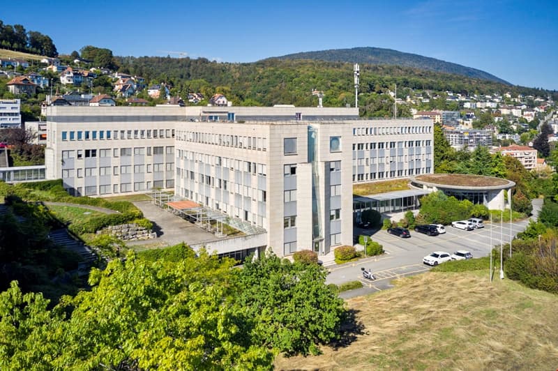 Modern office building with a green rooftop, glass facades, multiple floors, surrounded by a landscaped area, trees, and a parking lot with several cars.