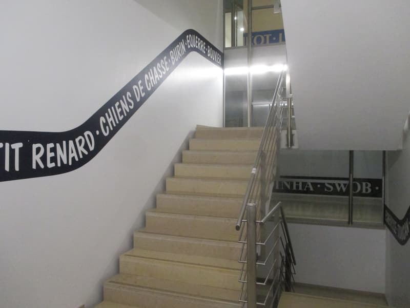 Inside image of a stairwell. It has stairs leading up and a white wall with a black written strip that says 'IT RENAR.' and some numbers. The railing of the stairs is silver.