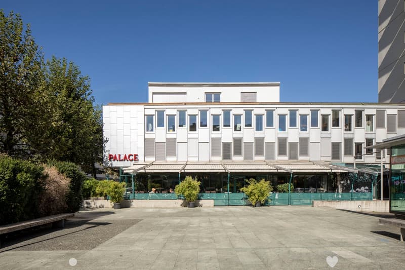 Modern, multi-story building with white exterior and rectangular windows, covered restaurant area, plants and benches in front