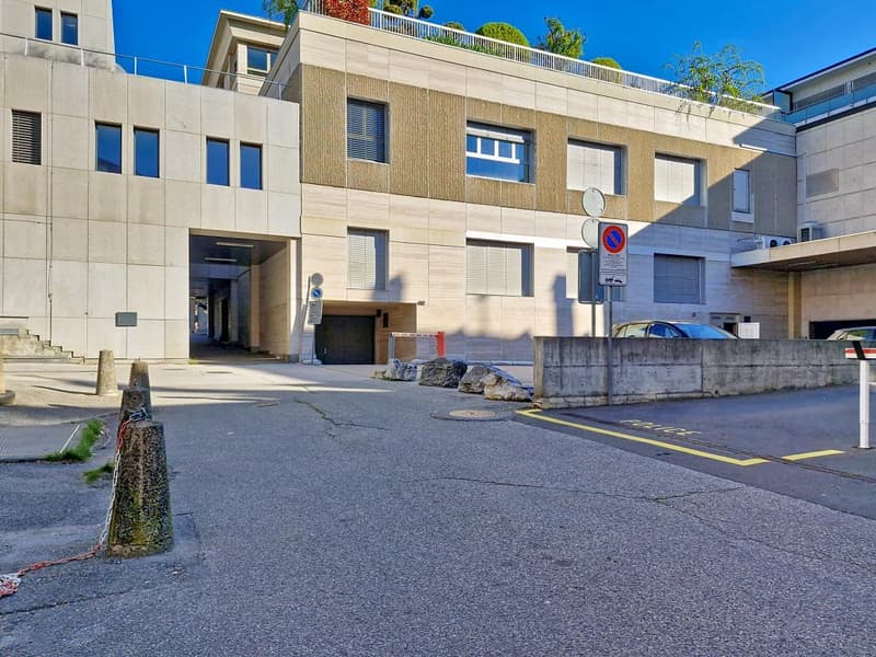 Modern building with multiple floors, concrete exterior, large windows, entrance with a gate, parking area, and a car parked in front.