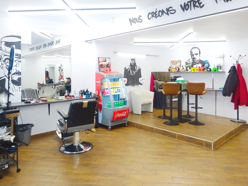 Barber shop with a counter, barber chair, refrigerator, stools, and a mirror. There are also some decorations on the wall.