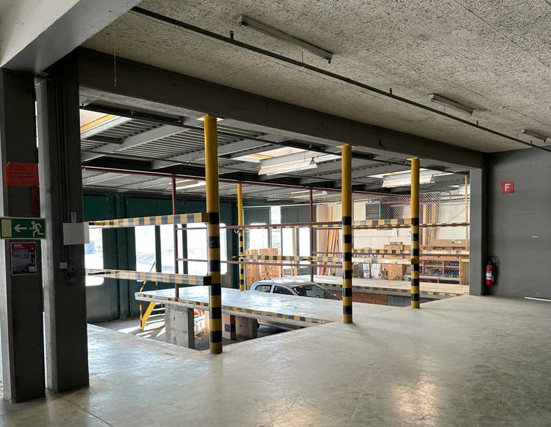 The image shows a commercial parking garage with concrete floors, yellow support columns, and storage racks along the walls. There is a car parked on the ground level, indicating this is a functional parking space.