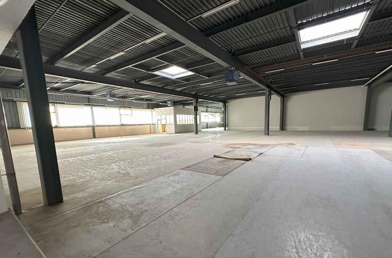 Large open space with concrete flooring, metal beams and ceiling, and natural lighting from windows. The space appears to be a warehouse or industrial facility.