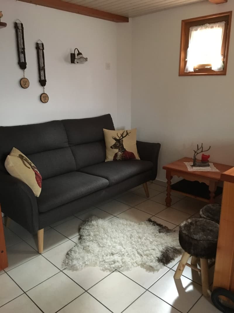 The image shows a cozy living room with a black sofa, a wooden side table, and a decorative pillow with a deer motif. The room has tile flooring and a white shag rug. There are wall decorations, including clocks and a deer-themed wall hanging. The room ha