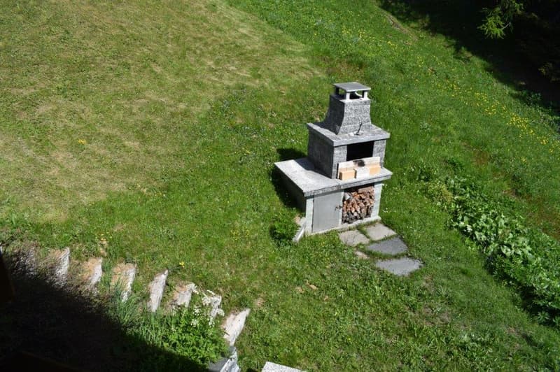 A stone outdoor fireplace with a chimney, situated on a grassy hill surrounded by greenery.