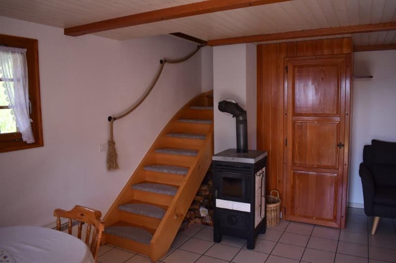 The image shows a cozy living room with wooden beams on the ceiling, a wooden staircase, and a wood-burning stove or fireplace in the corner. The room has a rustic, cabin-like feel with wooden paneling and a tiled floor.