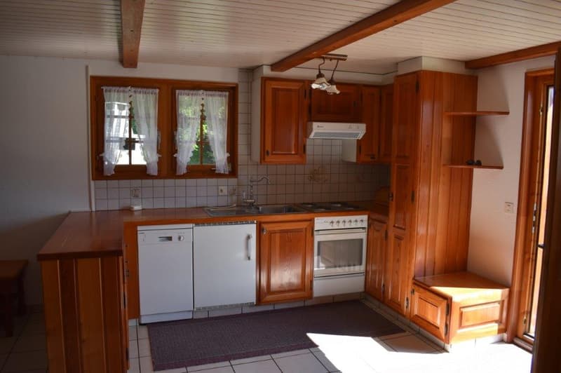 Spacious kitchen with wooden cabinets, tile countertops, stove, oven, and dishwasher. Large windows provide natural light. Tiled floor and exposed wooden beams on the ceiling.