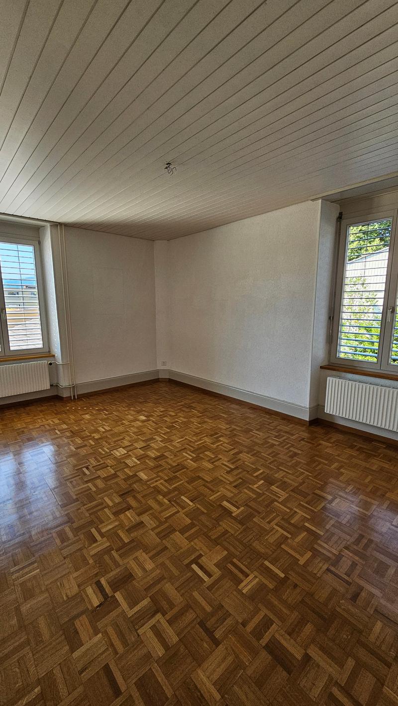 Wooden parquet floor, white walls, two windows with blinds, wooden radiators, ceiling beams