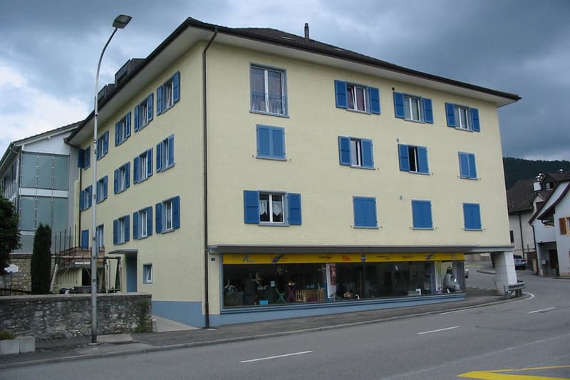 A multi-story yellow building with blue shutters on the windows. The building has a commercial space on the ground floor and multiple floors above. The building is situated on a street with a road and sidewalk in front of it.