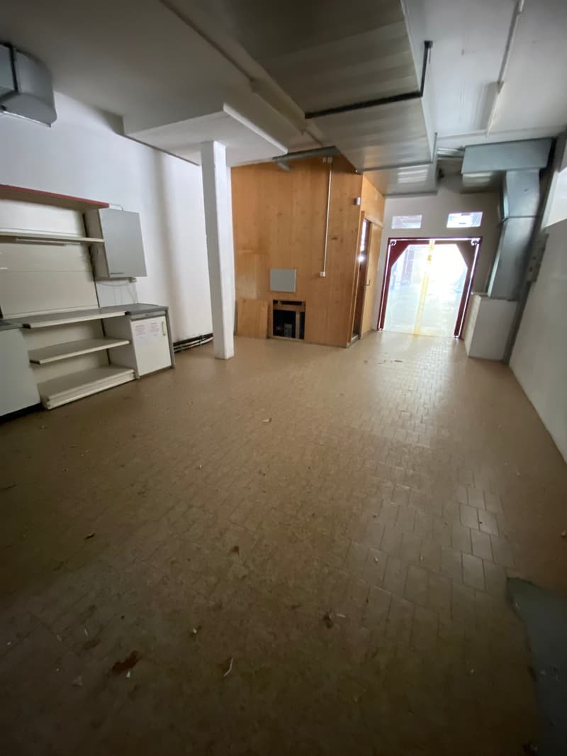 The image shows a large, open commercial space with a tiled floor. The space appears to be empty, with some shelving units and a wooden cabinet visible. The space has high ceilings and large windows, suggesting it could be used for a variety of commercial
