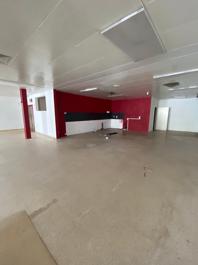 Large open space with concrete flooring, white ceiling, and red accent walls. The space appears to be in a commercial or office building setting, likely undergoing renovation or construction work.