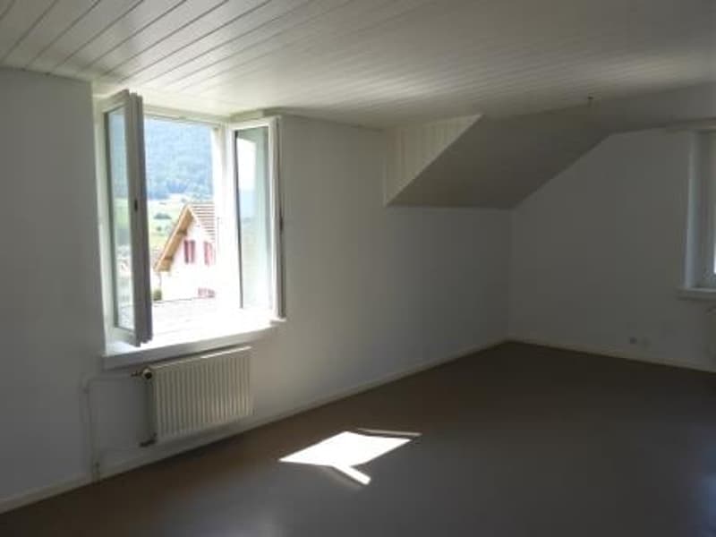 Spacious attic room with white walls, wooden ceiling, and a large window providing natural light. The room appears to be empty, with no furniture or decor visible.