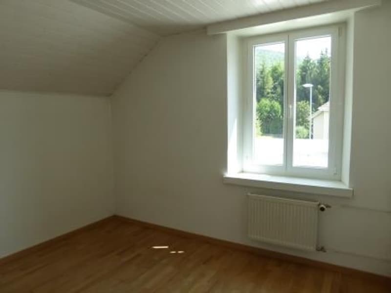Spacious bedroom with white walls, hardwood flooring, and a large window overlooking the outdoor greenery.