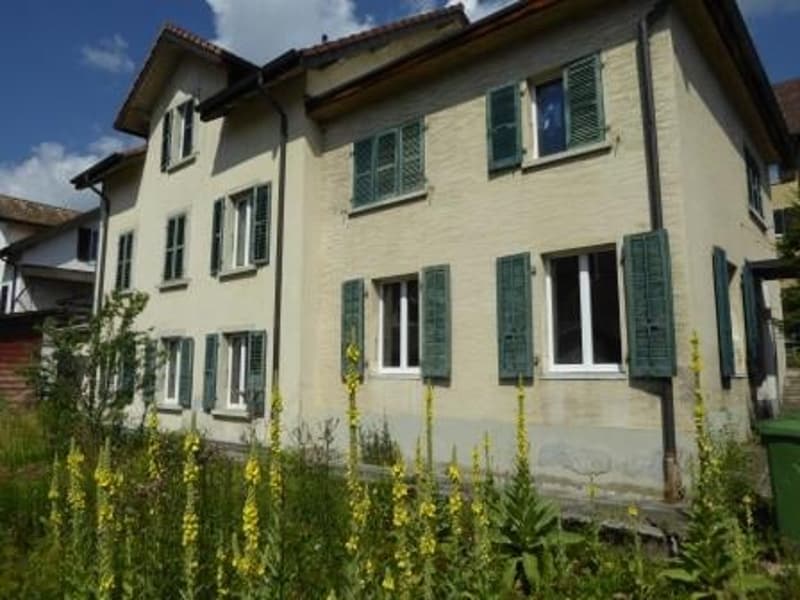 2-story house with yellow exterior, green shutters, and a balcony on the second floor. The house is surrounded by a grassy area with yellow flowers.