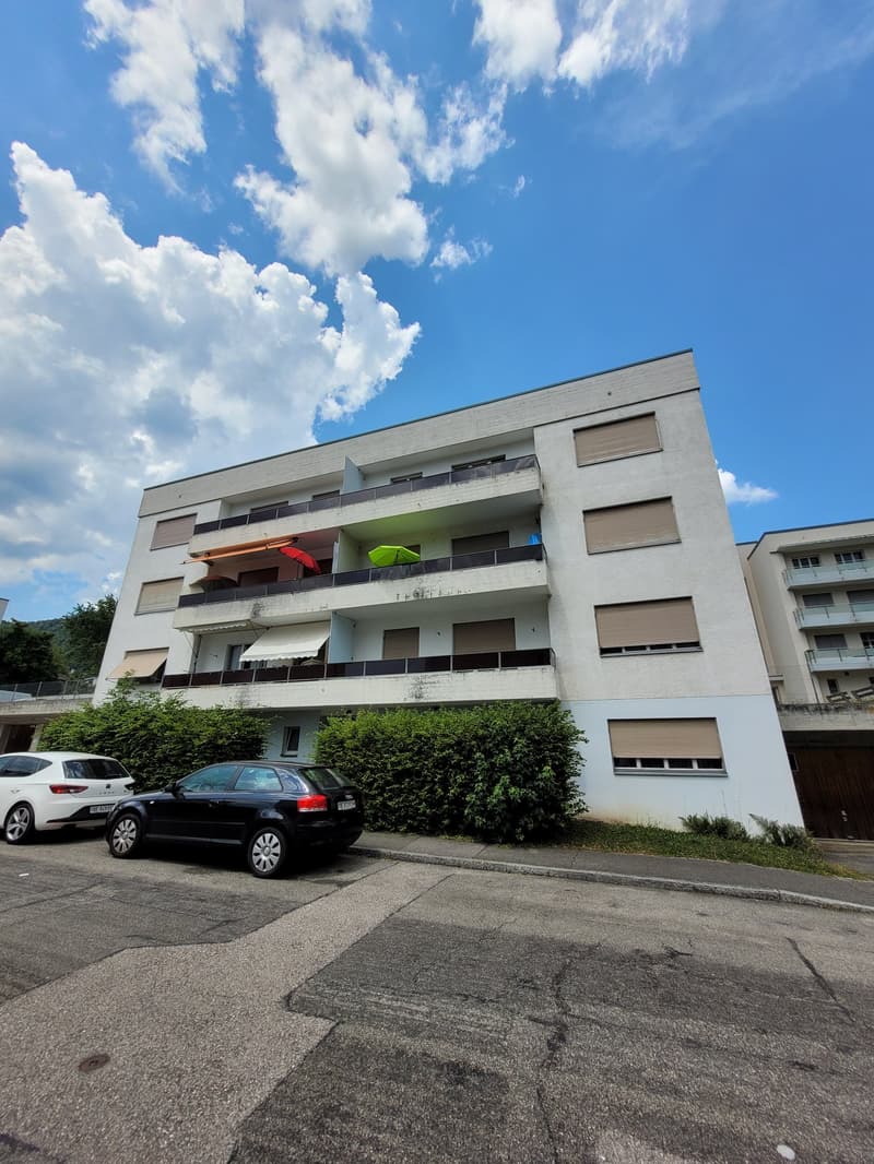 4-story apartment building, white walls, windows with brown shutters, plants in front, black car parked, another car in the background, blue sky with clouds