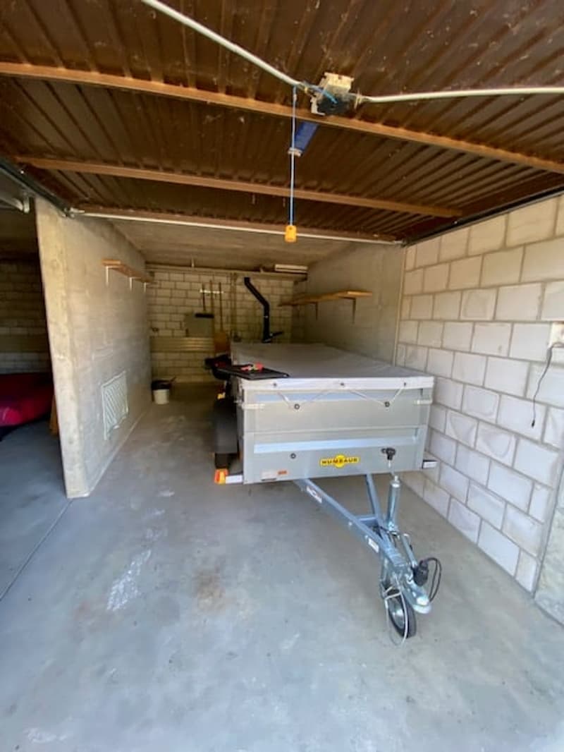 Empty garage with a trailer, brick walls, concrete floor, metal ceiling, open door, small bed, bucket, and shelves