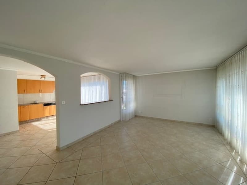 Empty room with white walls, beige tile flooring, windows with curtains, wooden cabinets, open space to a kitchen area