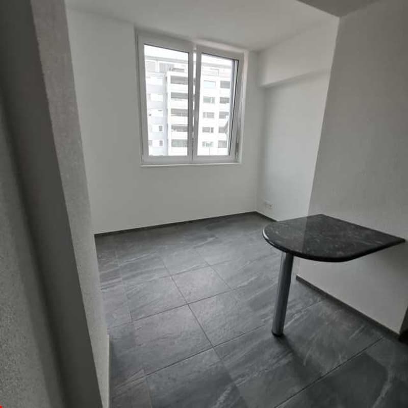 An empty room with tiled flooring, white walls, a corner table, and a window looking out at a building