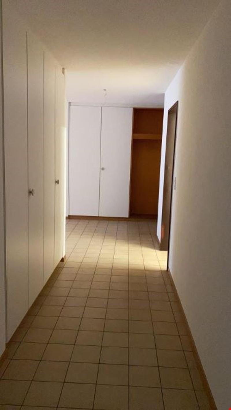 Empty corridor with white walls and tiled floor, three built-in cabinets on the left, a door in the center, and an open door leading to another room on the right.
