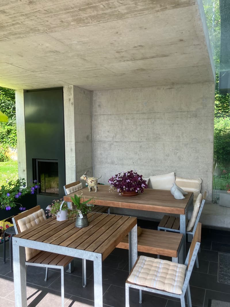 modern outdoor dining area with concrete walls, fireplace, wooden table, cushioned chairs, and various decorative items.