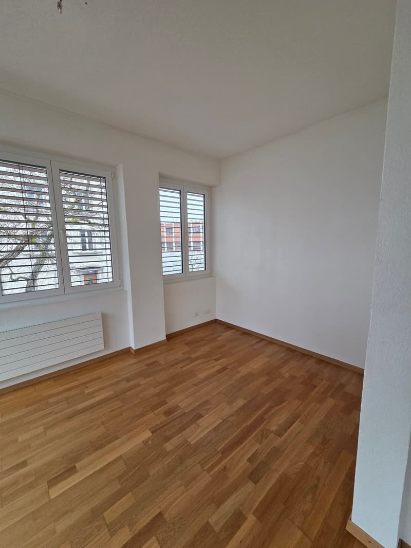 an empty room with hardwood floors, white walls, two large windows with blinds, and a radiator