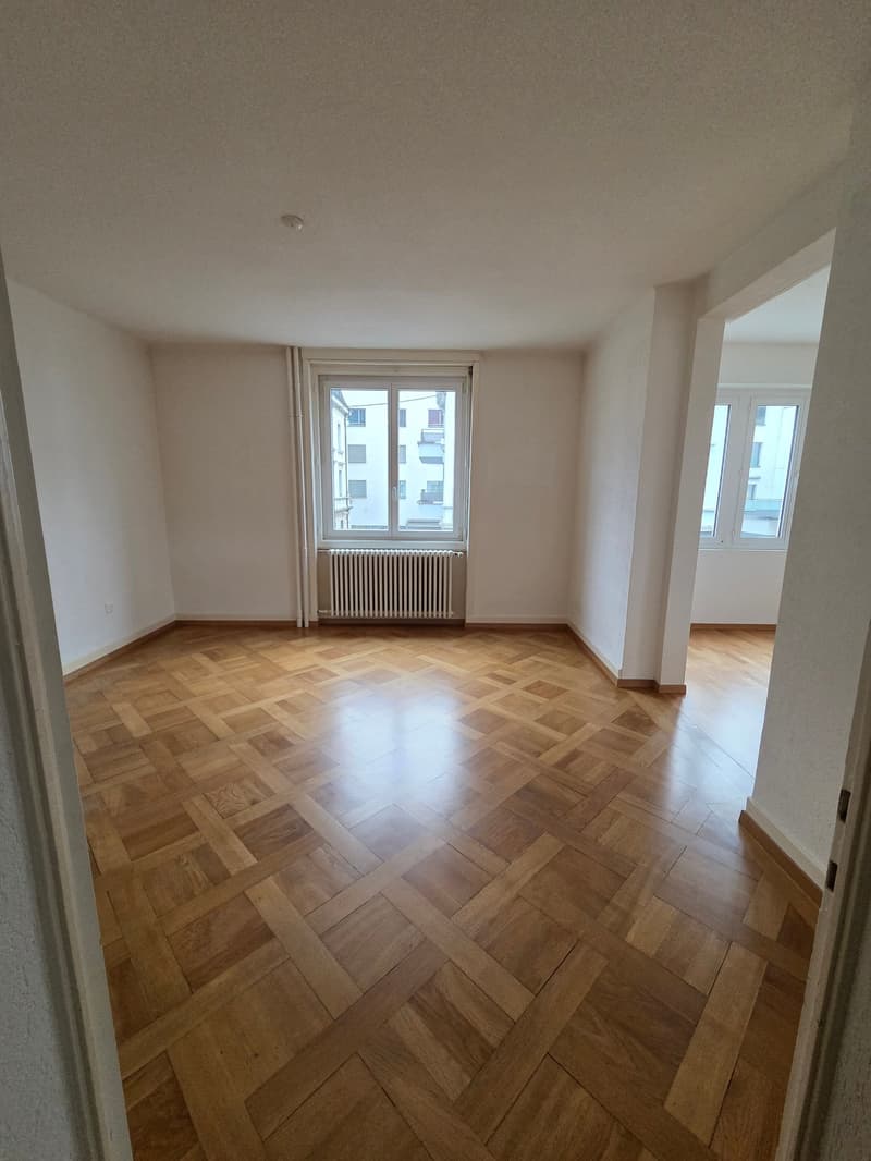 Spacious room with wooden parquet flooring, white walls, and large windows offering a city view
