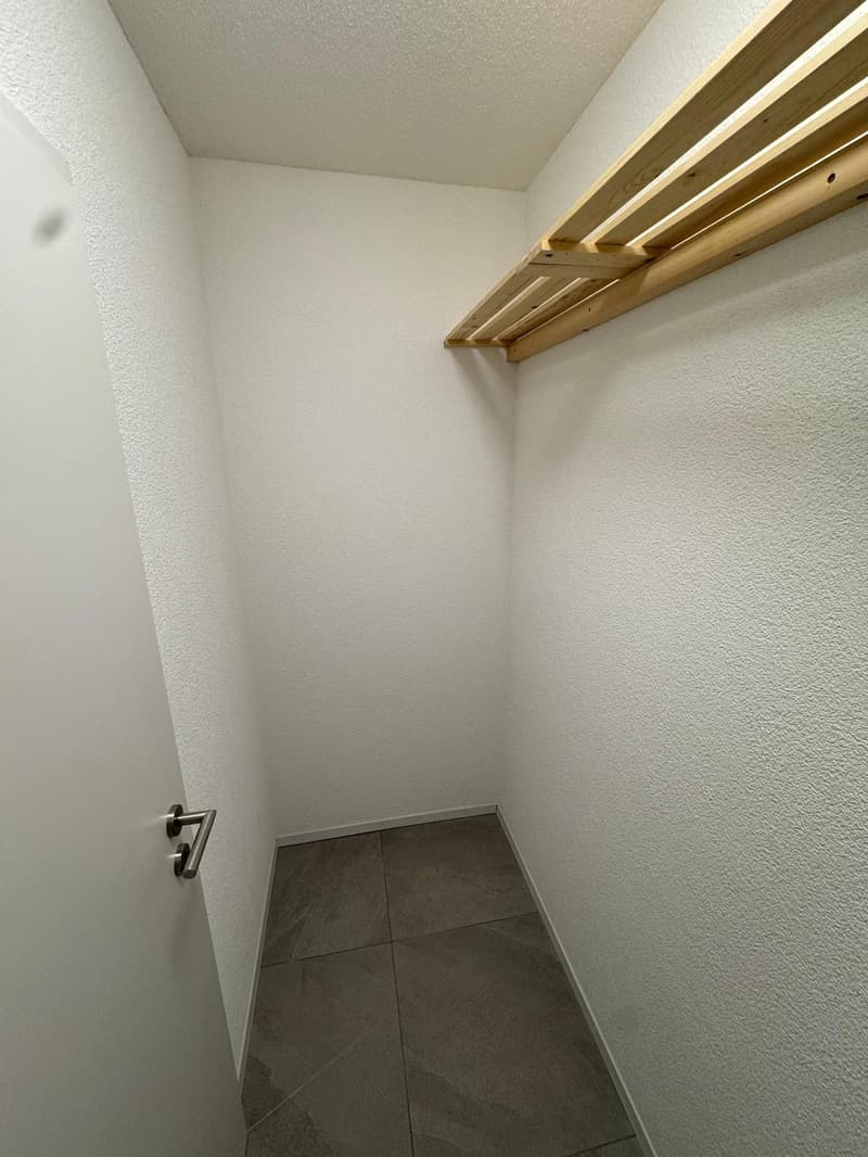 An empty room with white walls and a wooden shelf on the wall, floor tiles and an open door.