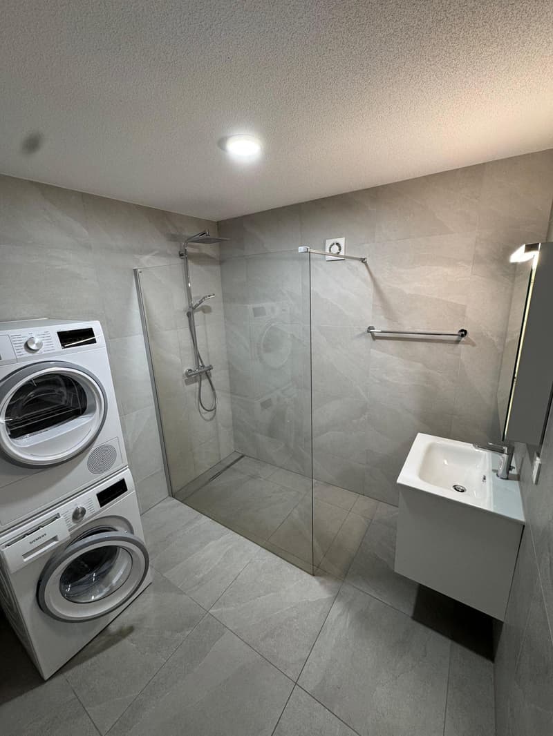 A modern and sleek bathroom with glass shower enclosure, white tiled flooring, washing machine, sink, and mirror, and a shower head