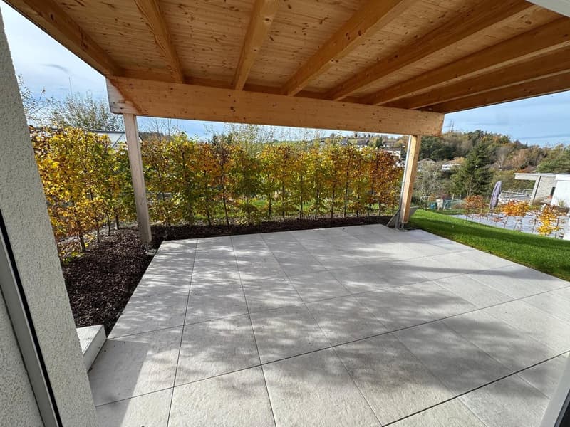 Outdoor area covered with wooden ceiling, tiled flooring, green grass, shrubs, bushes, trees, clear blue sky, small house in background