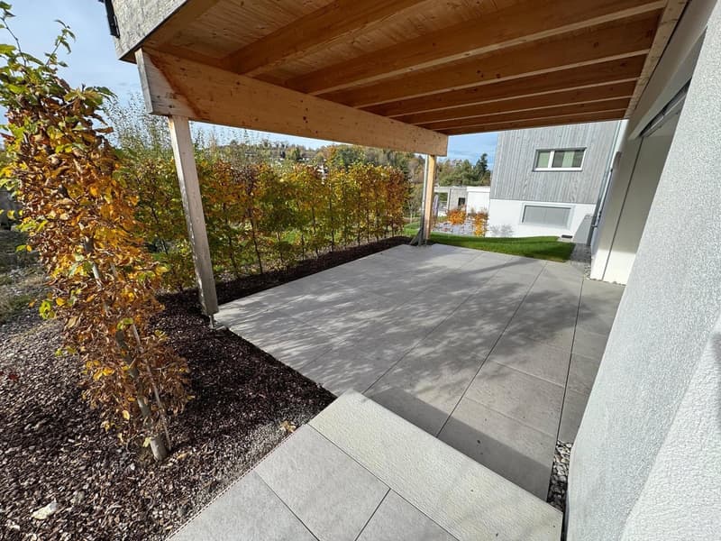Wooden pergola over a paved terrace, surrounded by greenery, adjacent to a modern house.