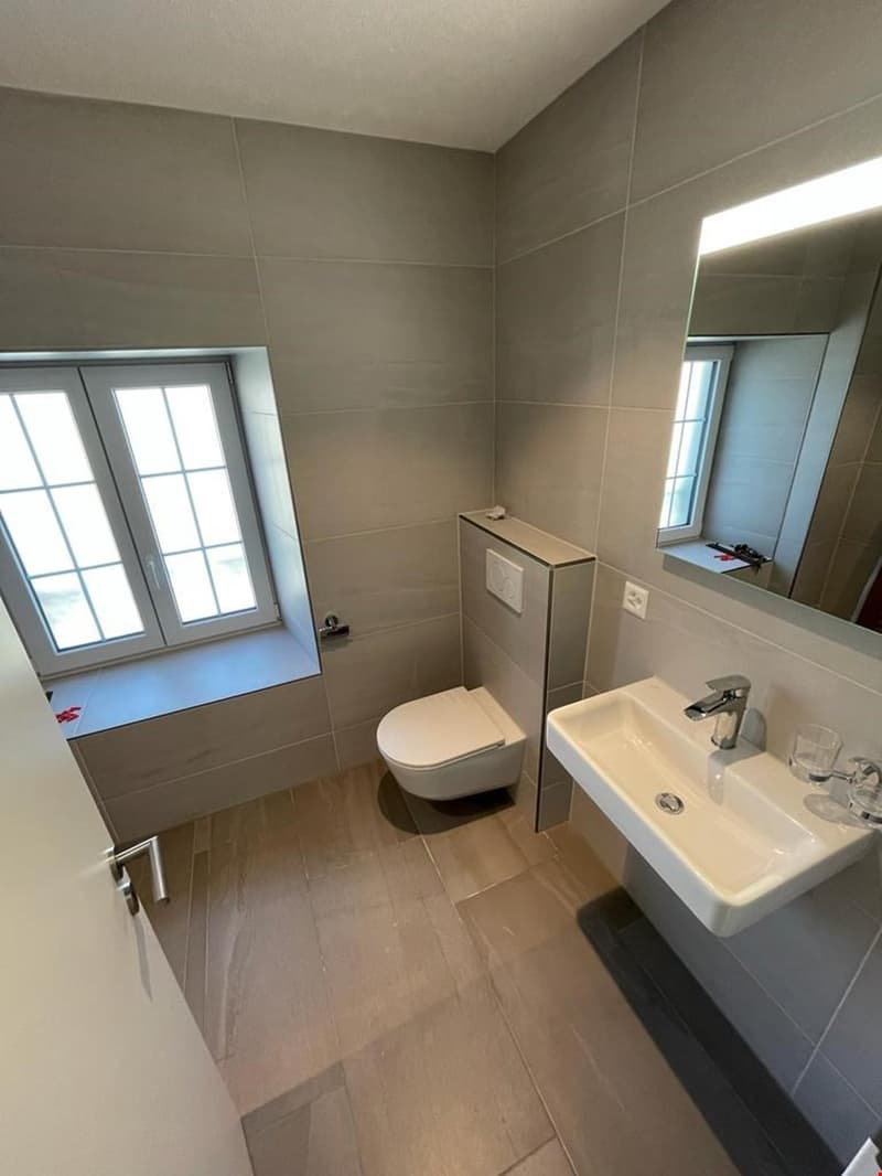 Modern bathroom with grey tiled walls, white sink, chrome faucet, white toilet, and large window