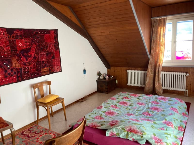 white wall, wood paneled ceiling, bed with floral cover, chair with pillow, window with curtains, small wooden desk, radiator, colorful tapestry