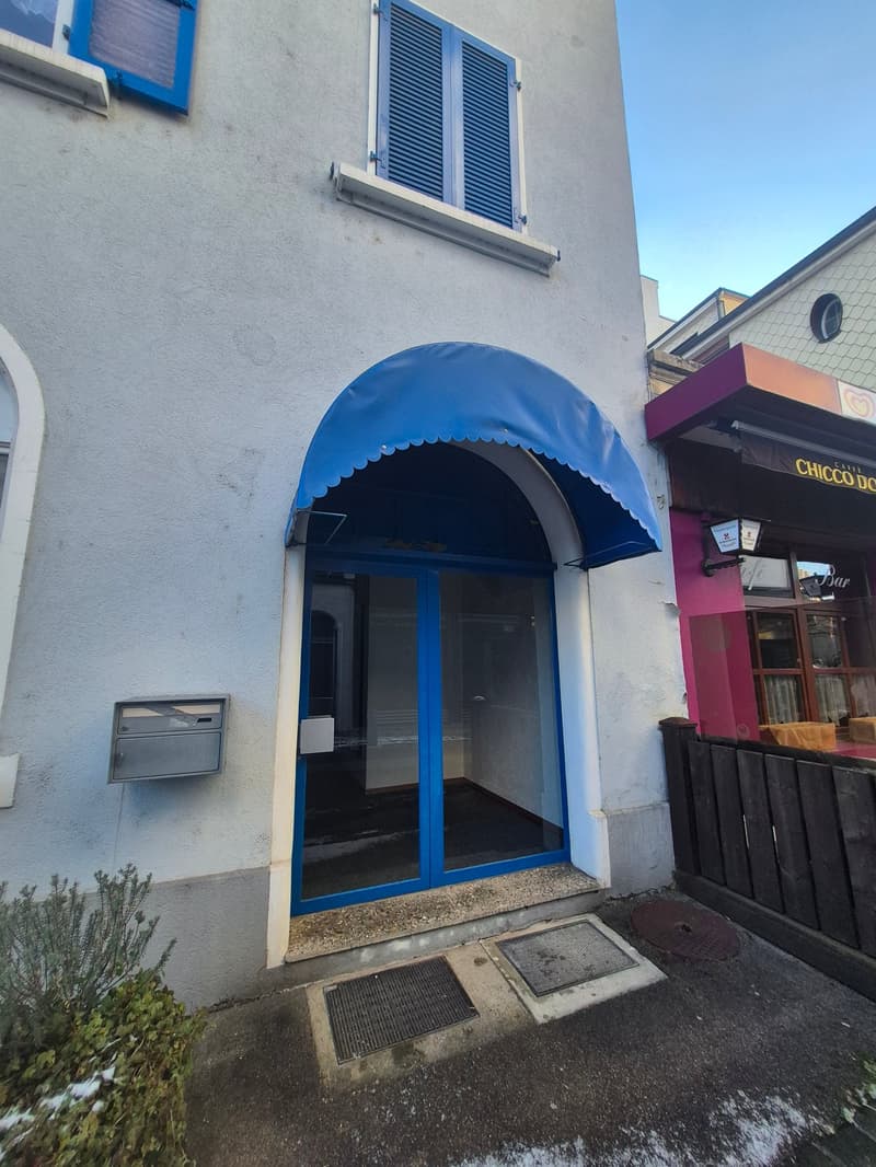 The entrance of the building has a blue awning over the entrance door, glass walls, and a glass window. There are plants on the side of the building. There are also other buildings on the side.