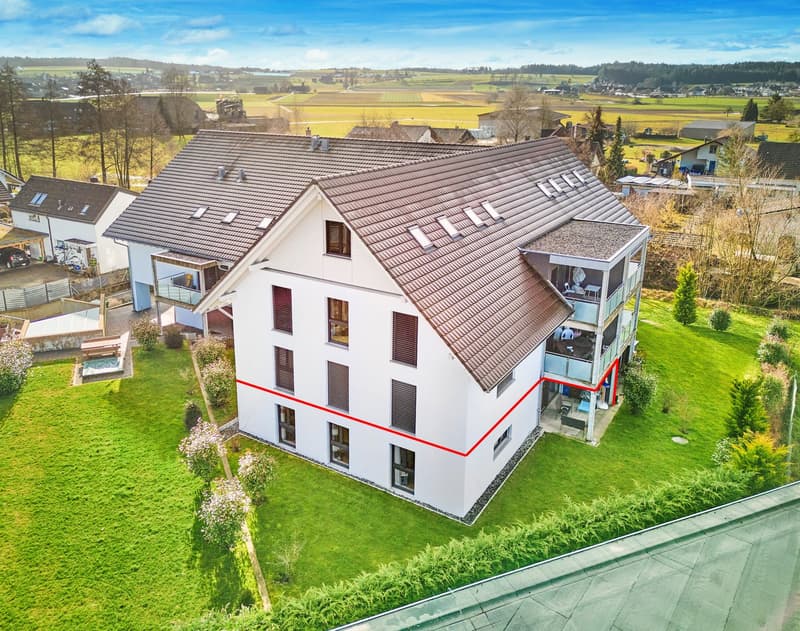 A two-story house with a brown roof and white walls, located in a grassy landscape with a field in the distance, balcony on the left side, garden on the right side.