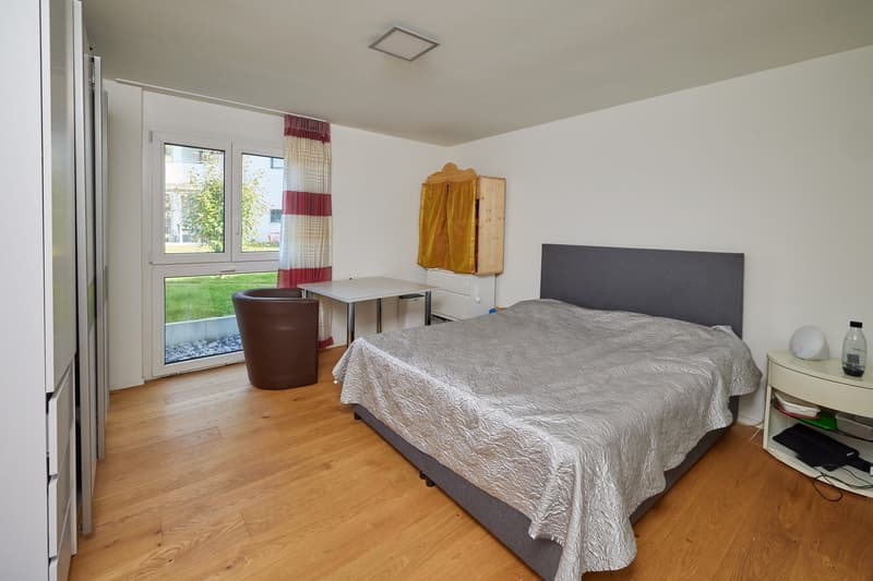 Bedroom with bed, desk, chair, wooden floor, white walls, window with curtains, small cabinet, and a view of the outside