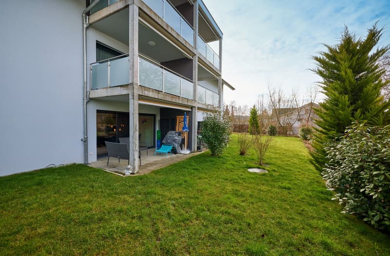 White building with balconies, garden, lawn, metal pole, green grass, trees, chairs, table