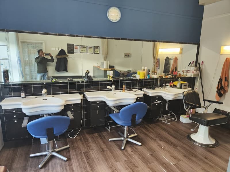 A room with a row of black cabinets with white basins, white wooden floors, and blue chairs, along with a mirror, clock, and a person taking a picture in the background.