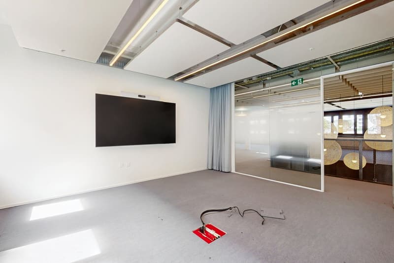 Large open office space with high ceilings, exposed beams, and a large TV screen on the wall. The room has glass walls and a fireplace visible in the corner.