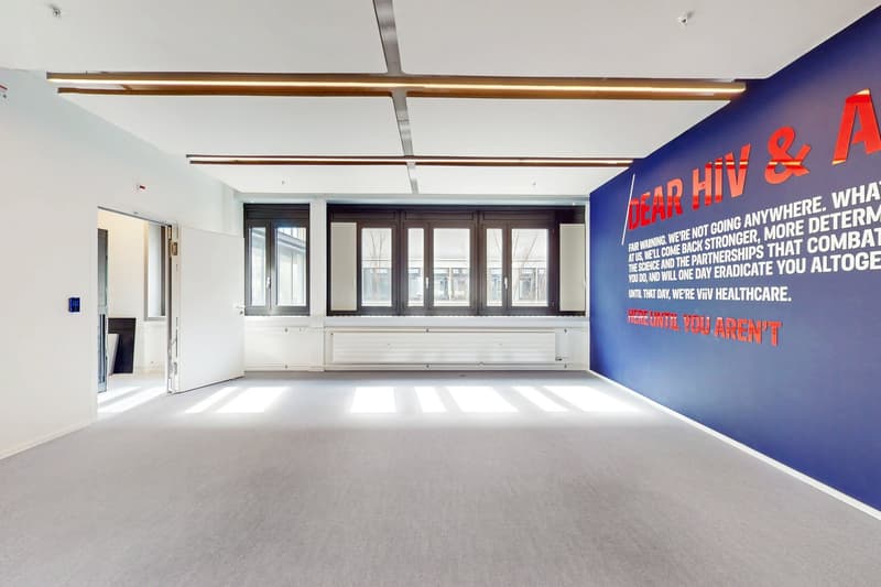 Large open room with white walls, gray flooring, and large windows. The room has a blue accent wall with a red and white text message about HIV and AIDS healthcare. The ceiling has long, horizontal lighting fixtures.