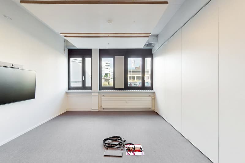 Large open room with white walls, hardwood ceiling beams, and large windows providing a view. The room is empty except for a TV mounted on the wall and some cables on the floor.