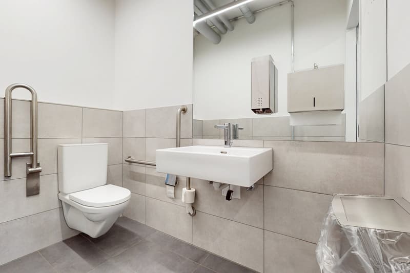 Modern bathroom with white toilet, sink, and tiled walls and floor. There is a dishwasher visible on the wall.