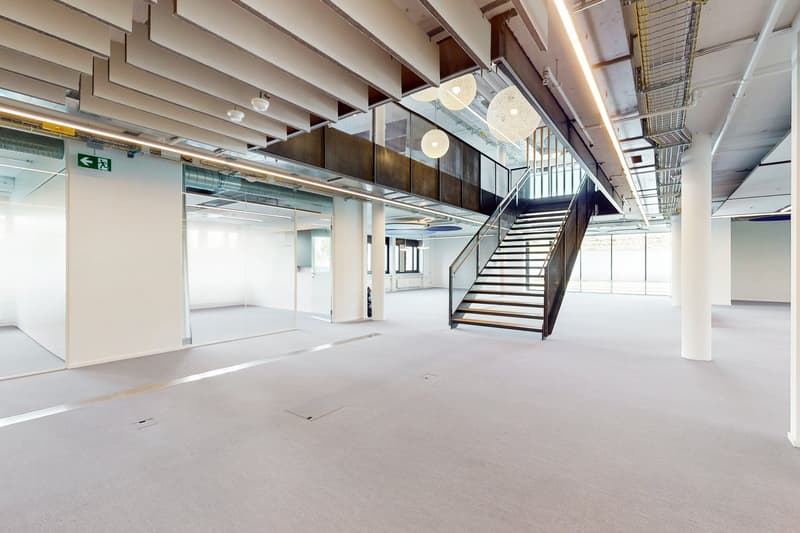 Large open-plan office space with high ceilings, exposed beams, and modern lighting fixtures. The space features a central staircase leading to a mezzanine level, and there are glass walls separating different areas. The floor is covered in a light-colore