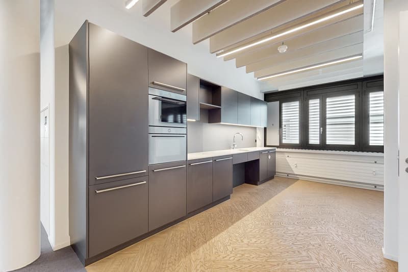 Modern kitchen with gray cabinets, stainless steel appliances including a dishwasher, and hardwood flooring. The kitchen has an open layout with large windows providing natural light.